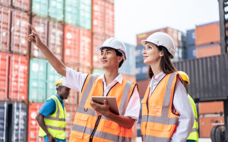 Caucasian business woman and Asian man working in container terminal. Attractive engineer people processes orders and product at warehouse logistic in cargo freight ship for import export in harbor.